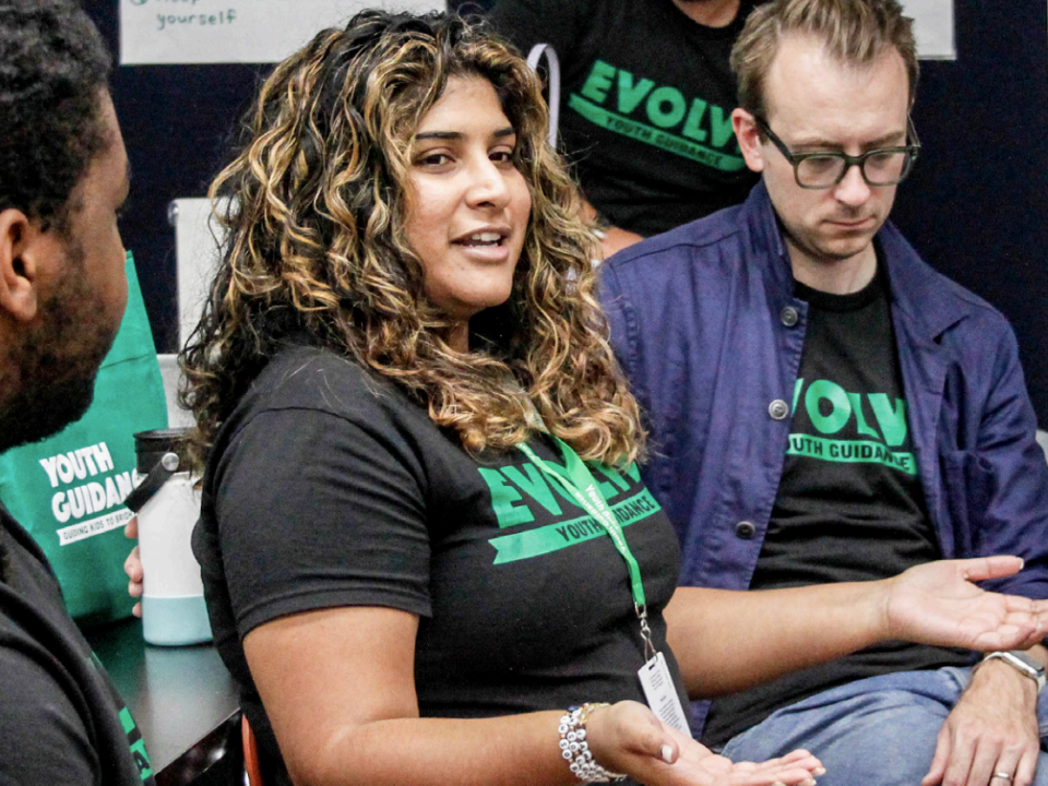 Three people sitting in a circle and talking to each other inside of a classroom. The woman has curly hair and is wearing a black t-shirt that says "Evolve." A man with glasses sits next to her, with his head down, listening intently. A man with short, dark beard stubble is seated on her right side and is making eye contact as she speaks.