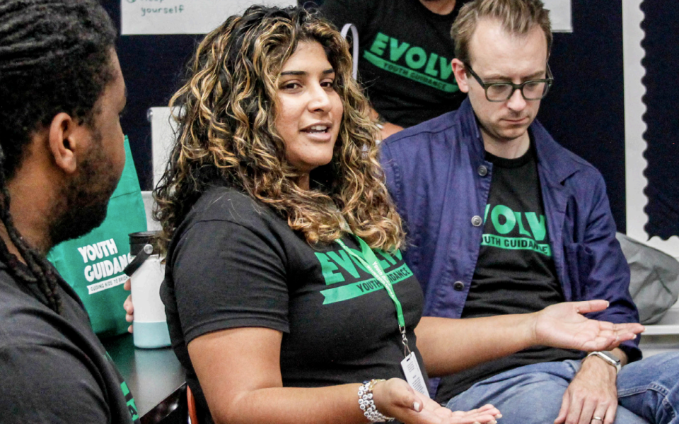 Three people sitting in a circle and talking to each other inside of a classroom. The woman has curly hair and is wearing a black t-shirt that says "Evolve." A man with glasses sits next to her, with his head down, listening intently. A man with short, dark beard stubble is seated on her right side and is making eye contact as she speaks.